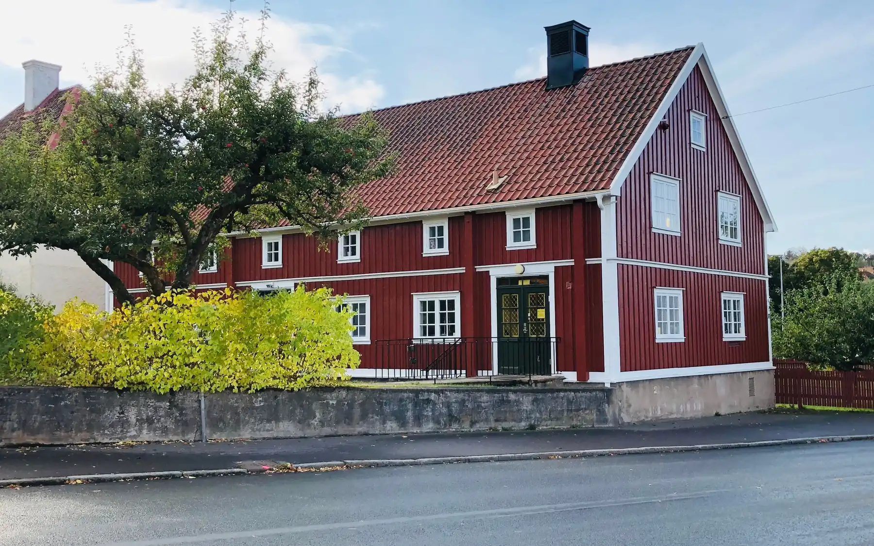 Langt rødt trehus med hvite vinduer og hage ved en gate